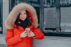 Pleasant looking young female in red jacket, holds modern smart phone, checks bank account, waits for call, enjoys online communication, stands outdoor, enjoys snowy weather. Spare time concept