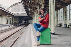 Pleased young woman has positive look, sits crossed legs at bag, holds cell phone, searches place on online map, makes video call, surfes internet website, waits for train at railway station photo