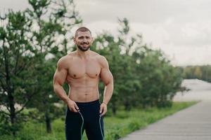 alegre deportista sin afeitar salta con cuerda, tiene cuerpo musculoso, disfruta del entrenamiento físico, posa al aire libre. hombre fuerte con torso desnudo usa equipo deportivo para hacer ejercicio, respira aire fresco en la naturaleza foto