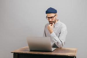 Image of unshaven male owner of trading company searches information about marketing agency, thinks about formal meeting, wears glasses and elegant clothes, sits at old desk over grey background photo