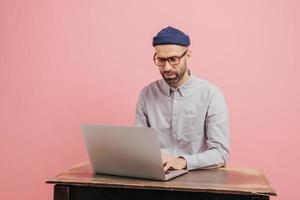 Concentrated hipster student reads information and checks data, focused in monitor of laptop computer, keyboards something, sits at desk, wears formal clothes, isolated over pink studio wall photo