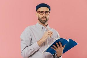 Isolated shot of attractive young male holds textbook, makes notes in organizer, dressed formally. Clever student prepares for examination or does home assignment, isolated over pink studio wall photo