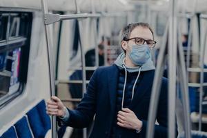 Serious European man being aware of his health, protects from catching serious disease in public transport, wears medical mask on face, travels in urban train, afraids of coronavirus epidemy photo
