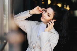 Elegant brunette slim woman with pony tail dressed in white coat having dark eyebrows, eyes, pure skin, nice manicure chatting over mobile phone touching her head with hand looking smiling aside photo