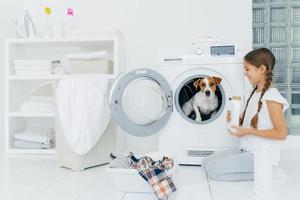 Cheerful little girl stands on knees with washing detergent, poses near washing machine, looks happily at favourite pet inside of washer, going to do laundry at home, spends free time with dog photo