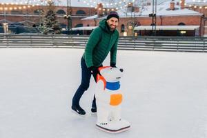 Unexperienced male going to learn skating skills, uses skate aid for being in balance on ice rink and not fall, expresses positive emotions and enjoyment, rejoices winter holidays. Entertainment photo