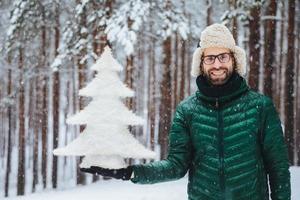 personas, emociones, clima nevado y concepto de temporada. feliz y alegre joven sin afeitar vestido con ropa de abrigo, sostiene un abeto, se para contra el fondo de los árboles, disfruta de hermosos paisajes naturales foto