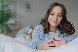 Indoor shot of attractive thoughtful young woman lies in bed, uses modern cell phone, watches video or film online, enjoys good sound, dressed in pyjamas. People, bed time and relaxation concept photo