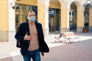 European man wears protective face mask while coronavirus and pandemic, carries rucksack, poses outdoor at bus station, travels during quarantine. Covid-19 virus, epidemic outbreak, public place