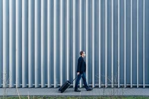 Horizontal panoramic shot of man passenger arrives in own country because of quarantine and pandemic situation in world, walks with suitcase, poses outdoor against metal fence, wears face mask photo