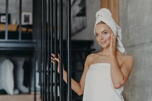 Beautiful healthy young woman applies face cream on complexion, looks thoughtfully somewhere into distance, wrapped in towels, stands indoor, has perfect pure skin. Women and cosmetology concept photo