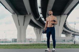 Muscular athletic bearded bodybuilder raises dumbbells works on biceps has hard workout near bridge dressed in sport trousers and sneakers, being motivated, enjoys sport. Healthy lifestyle concept
