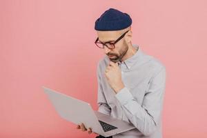 Experienced male freelancer develops new strategy, holds modern laptop computer, keeps hand on chin, wears formal clothing, busy with distant work, isolated over pink background, copy space. photo
