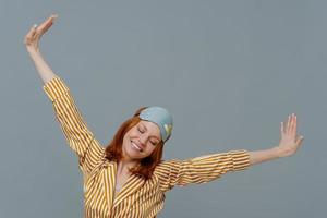 Perfect rest and sleeping concept. Cheerful young ginger European woman spreads arms and stretches after awakening, had sweet dreams, wears striped pajama and sleepmask, isolated on grey wall photo