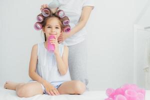 Cropped shot of pleased little child with curlers on head and her mother stands behind combs hair and winds curlers, have appealing appearnce. People, loving family and relationship concept. photo