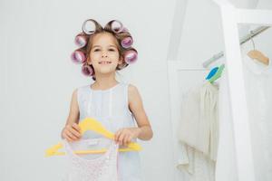 Indoor shot of fashionable cute small girl with gentle warm smile, prepares for something, has curlers on hair, holds new outfit on hangers, look joyfully at camera. Children and beauty concept photo
