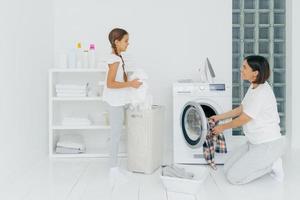 contenta de que la madre y la hija estén ocupadas lavando la ropa en casa, tienen caras felices, la mujer morena carga la lavadora, la niña se para cerca de la canasta con ropa blanca. tareas domésticas, concepto de limpieza. foto