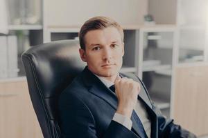 Attractive male manager sits in chair at his cabinet, dressed formally, rests for minute after hard work in office. Prosperous man boss has serious expression, works on financial report. Business photo