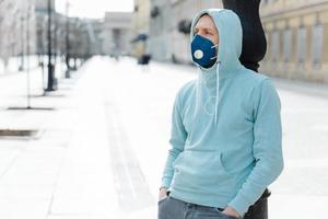 Outdoor shot of hipster wears sweatshirt and respiratory mask, walks in epidemic city, prevents virus, infection and coronavirus, afraids of being infected. Qaurantine time. Health safety concept photo