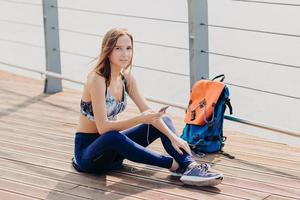 mujer encantadora relajada con cabello oscuro, piel sana y pura, vestida con ropa deportiva, sostiene el teléfono celular para comunicarse, escucha su canción favorita, la mochila está cerca de ella. gente, deporte, concepto de recreación. foto