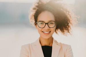 Headshot of cheerful curly woman with positive expression, wears spectacles, white elegant jacket, looks straightly at camera, stands outdoor, has curly hair expresses good emotions. Ethnicity concept