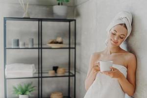 una mujer europea sana y satisfecha posa cerca de la pared en el baño, envuelta en toallas blancas y suaves, sostiene una taza de té, se relaja después del tratamiento de spa y se baña, disfruta de los tratamientos de higiene en casa