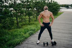 Bearded muscular man keeps hands on waist, raises head and breathes fresh air, takes break after doing workout, trains with barbells. Professional weightlifter works out in nature. Exercising concept photo
