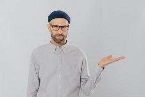 Indoor shot of unshaven Caucasian man holds copy space, demonstrates something against white wall, involved in advertising campaign. Serious male model with bristle stands alone. Isolated shot photo