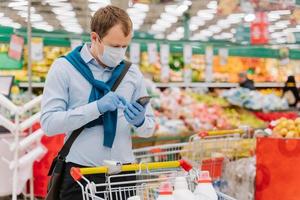 Young man poses in grocery store during coronavirus pandemic, wears protective medical mask and gloves, stands in supermarket near trolley, checks something in smartphone. Health, safety, quarantine