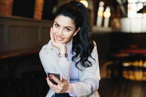 Indoor shot of brunette female having dark eyes with long eyelashes, full well-shaped lips holding her hand on cheek listening to music with headphones and smartphone having pleased expression photo