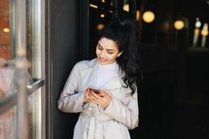 Portrait of fashionable brunette woman with pleasant appearance having nice manicure wearing white raincoat holding smartphone in her hands having pleasant smile being glad to recieve message photo