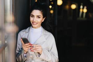 Sexy brunette lady with appealing dark eyes and full lips having red manicure holding cell phone while communicating with her friends having pleasant smile while looking into camera. Fashion concept photo