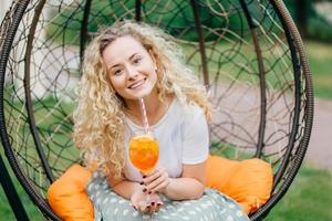 Photo of attractive beautiful curly light haired female with cheerful expression, recreats outdoor in hanging chair, drinks cocktail enjoys sunny day during summer. People, rest and lifestyle concept.