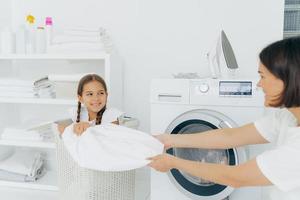 Happy girl with two pigtails poses in basket with dirty linen, has fun in laundry room with mother, helps to do washing. Woman loads washing machine, spend weekend at home, busy with domestic work photo