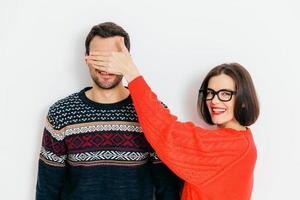 Portrait of happy covers eyes to her husband, prepares surprise for him, wears casual red oversized sweater, has positive smile, isolated over white background. I am going to show something to you photo