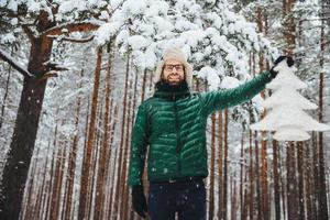 toma horizontal de un atractivo hombre barbudo que se alza contra los árboles de invierno en el bosque, sostiene un abeto artificial blanco, expresa un estado de ánimo festivo y se prepara para las vacaciones de invierno. gente, clima, temporada foto