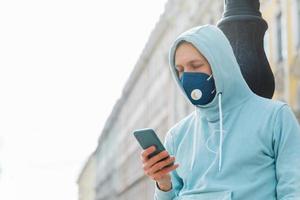 Man on street wears respiratory mask, walks through city, uses modern mobile phone concentrated into display uses wireless internet prevents himself from coronavirus infection. Rapidly spreading virus photo