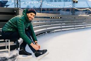 Cheerful man with appealing appearance laces up skates, sitts on ice arena, wants to go skating with friends, poses at camera. Happy bearded man enjoys having active lifestyle. Winter and season photo
