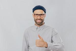 Positive smiling man with stubble, raises thumb up, demonstrates his like and approvement, wears headgear and formal shirt, isolated over white background. My answer yes. Gesturing. Body language photo