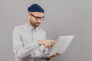 Professional male freelancer makes project work, points with index finger at screen of laptop computer, watches training webinar, holds modern gadget on hands, stands against grey studio wall photo