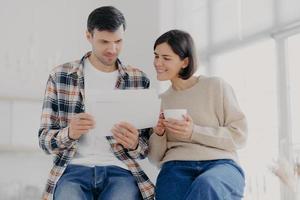 Indoor shot of happy wife and husband study documents received from bank, do paperwork together, drinks aromatic coffee, stand closely to each other, dressed in casual wear, pose in cabinet. photo