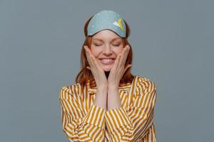 Pleased woman touches cheeks, has healthy freckled skin, smiles broadly prepares for sleep, wears striped pajama and sleep mask, closes eyes, isolated over grey background. Rest and lifestyle concept photo