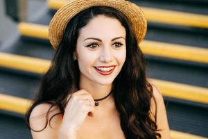 Pretty woman with dark hair, eyes, pure skin and red lips wearing straw hat looking with enjoyment into distance while sitting at stairs. Fashionable female in summer hat showing her emotions photo