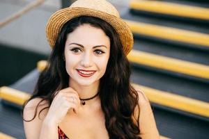 Outdoor portrait of fashionable woman with dark thick hair having make-up and painted red lips wearing summer straw hat and necklace looking into distance having gentle smile sitting at stairs photo