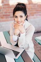 mujer morena lujosa con una sonrisa encantadora que mira agradablemente a la cámara mientras se sienta en el café frente a una computadora portátil abierta charlando con amigos o su novio. concepto de personas y tecnología foto