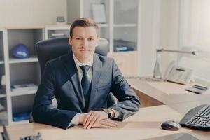 Indoor shot of pleasant looking male manager in black suit, sits in office, has confident expression at camera, works during working day, makes decision concerning future plans. CEO in cabinet photo