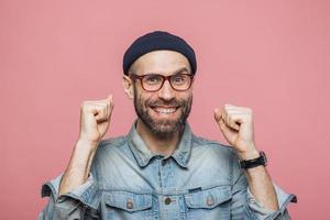 feliz hombre barbudo con ojos azules y expresión alegre aprieta los puños, se siente ganador, demuestra positividad, aislado sobre fondo rosa. emocionado emocional joven posa interior foto