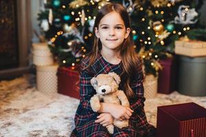 Small female child with appealing appearance, holds teddy bear, recieves present from parents, sits near Christmas tree. Adorable girl being glad to get gift on New Year. Christmas time. photo