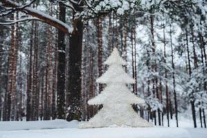 Horizontal shot of artificial white New Year tree stands in winter forest against natural trees. Outdoor photo of Christmas white tree on beautiful nature