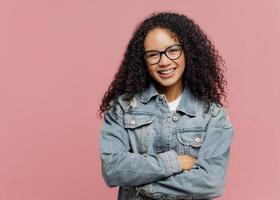 Horizontal shot of mirthful pleasant looking female model wears optical glasses and jean jacket, keeps arms folded over chest, enjoys spare time, nice friendly conversation, poses indoor on pink wall photo